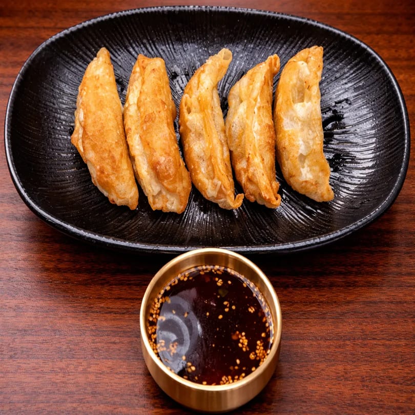 Five golden-brown fried dumplings on a black plate, with a small bowl of soy sauce and sesame seeds.