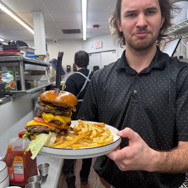 A man in a kitchen holds a giant burger with fries, looking directly at the camera.