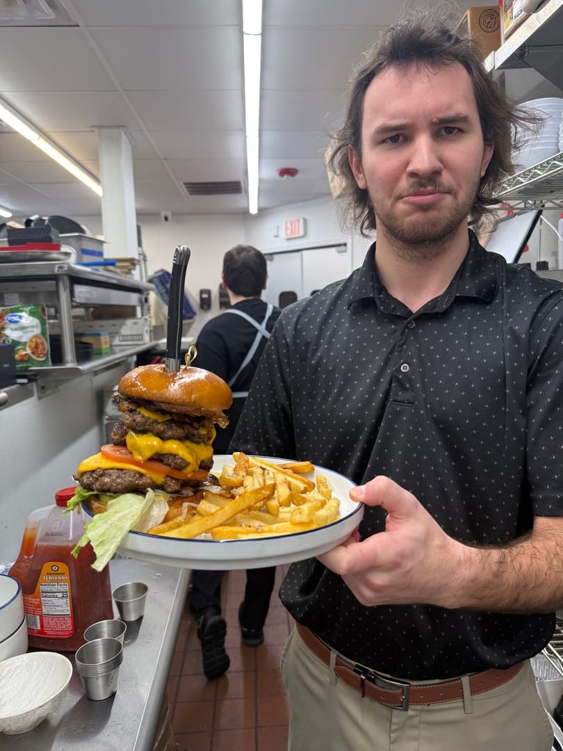 A man in a kitchen holds a giant burger with fries, looking directly at the camera.