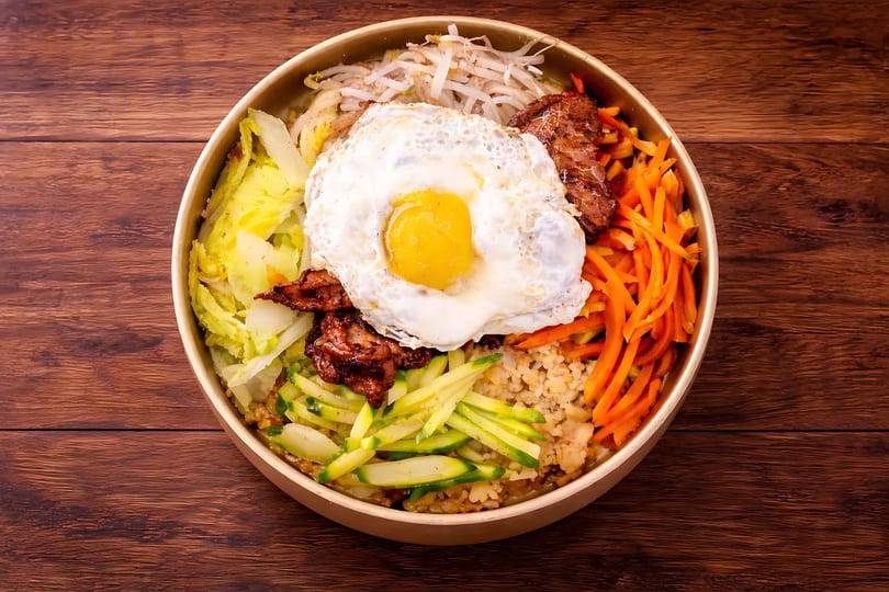Bibimbap bowl with rice, beef, fried egg, carrots, zucchini, bean sprouts, and cabbage.