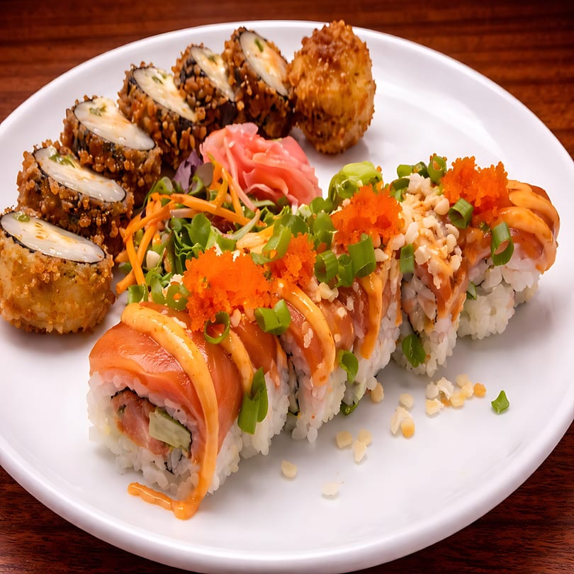 A white plate holds various sushi rolls, including salmon, fried, and roe-topped varieties, with ginger and greens.