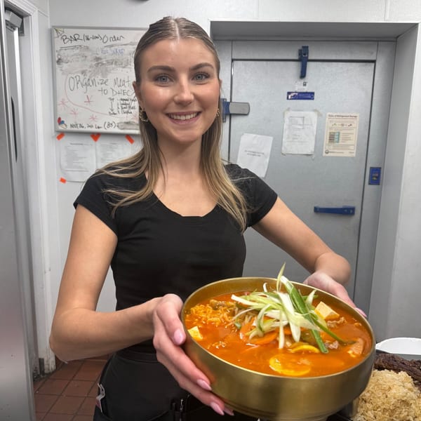 Smiling server holding a large golden bowl of Korean stew with scallions, tofu, and meat.