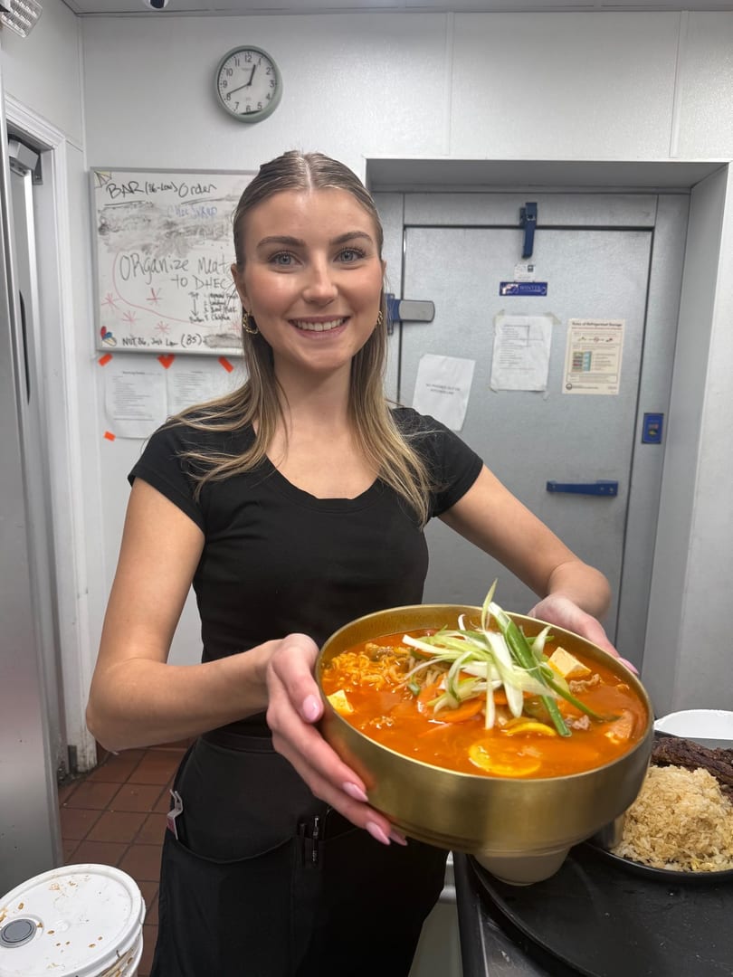 Smiling server holding a large golden bowl of Korean stew with scallions, tofu, and meat.